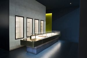 A modern, minimalist jewelry store interior with a long glass display counter containing jewelry pieces, set against dark blue walls and a lit display wall with empty glass shelves.