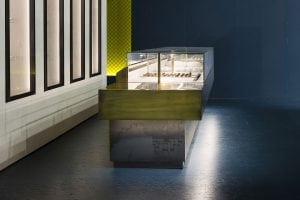 A modern, minimalist pastry display case with a metallic finish holds several desserts. The case is lit from above, set against dark blue walls and a yellow mesh accent in the background. The floor is a smooth, dark surface.