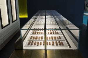 A modern glass display case showcases neatly arranged rows of small chocolate pastries on trays, with white pastries displayed on the upper shelf in the background.