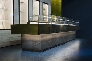 A modern, empty display counter with glass panels and a metallic finish stands in a minimalist interior, with shelves visible in the background and soft lighting highlighting the surfaces.