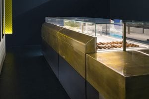 A modern dessert display case with a sleek brass finish holds trays of pastries, including a row of chocolate eclairs, inside a dimly lit, contemporary bakery interior.