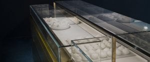 A glass display case filled with various types of white cheese, including mozzarella balls and cheese wheels, arranged on plates and trays. The case has a metal frame and sits against a dark background.