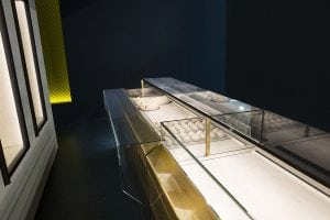 A modern, empty glass display case with a gold frame sits in a dimly lit room. One section contains several white ceramic bowls, while the rest of the case is empty. A yellow-lit mesh wall is visible in the background.