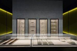 A modern, minimalist bakery display with glass cases showing assorted pastries and chocolates. Behind the counter are shelves with white cakes and desserts, set against a gray wall with yellow lighting accents.
