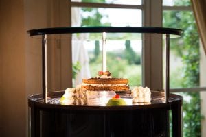 A lit birthday candle stands on a cake displayed on a round glass cake stand, surrounded by meringues and a small green pastry, with sunlight streaming through a window in the background.