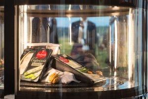 Several vacuum-sealed packages of cheese and a few walnuts are displayed on a rotating shelf inside a glass refrigerator case, with reflections and blurred figures visible in the background.