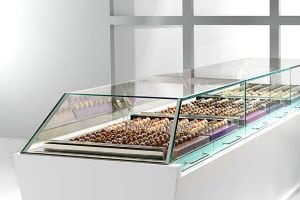 A modern glass display case filled with rows of assorted chocolates and truffles, arranged neatly in trays, set in a clean, bright environment with a grid-patterned wall in the background.