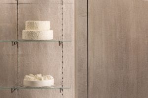 Two glass shelves display cakes with white frosting; the top shelf holds a two-tiered cake with textured icing, while the bottom shelf holds a single-tier cake decorated with white frosting roses. The background is beige.