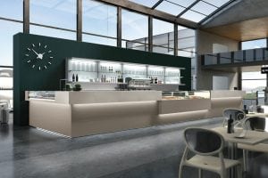 A modern, minimalist café counter with display cases and glass shelves, set in a bright space with large windows, a dark green accent wall, and a large wall clock; tables and chairs are arranged in the foreground.