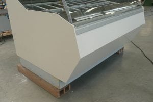 A large, white refrigerated display case sits on a wooden pallet in an industrial setting with a concrete floor. The case has a glass top and angled front, and appears to be newly manufactured or awaiting installation.