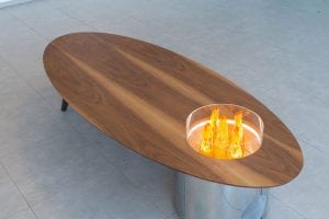 An oval wooden coffee table with a circular glass section containing decorative orange crystal-like sculptures, placed on a light-colored tiled floor.