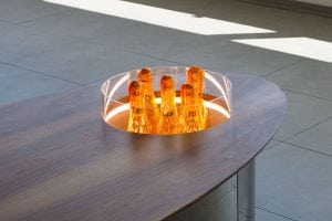 A modern oval wooden table with a round inset featuring illuminated amber-colored glass sculptures encased in a clear cylinder. The table is in a sunlit room with tiled flooring.