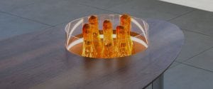 A modern wooden table with an embedded circular glass display containing several upright, amber-colored crystal-like sculptures. The display is illuminated, highlighting the bright orange pieces.