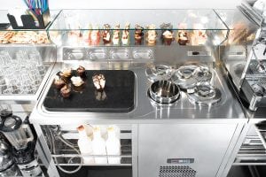 A stainless steel dessert station with cupcakes, ice cream or gelato in covered tubs, glassware, toppings in trays, bottles of sauces, and assorted dessert toppings displayed behind a glass sneeze guard.