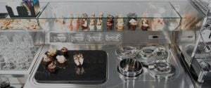 A display case in a modern dessert shop with an assortment of cupcakes and pastries arranged behind glass, with glasses and dessert-making equipment on the stainless steel counter.