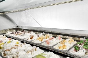 A display case filled with various flavors of gelato, mostly in white or light colors, topped with different decorations like fruits, chocolate, and cookies. The gelato is arranged in metal trays under a glass cover.