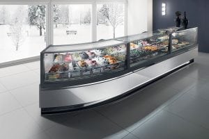 A modern glass display case filled with assorted gelato and desserts stands in a bright, sleek room with large windows showing snowy trees and a winter landscape outside.