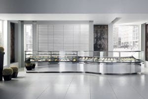 A modern, minimalist bakery or café with large floor tiles, tall windows, and sleek glass display cases filled with pastries and desserts. Decorative potted cacti are placed in the bright, spacious interior.