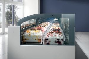 A modern gelato display case filled with multiple trays of colorful, assorted gelato flavors and toppings, positioned in a bright, minimalistic room with large windows.