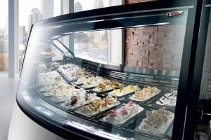 A curved glass display case filled with multiple trays of colorful gelato and ice cream toppings, set in a modern shop with large windows overlooking a city skyline.