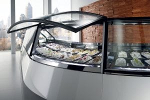 A sleek, modern gelato display case filled with various colorful gelato flavors stands in a bright room with large windows and city buildings visible outside.