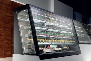 A glass display case filled with a variety of cakes and desserts, arranged on multiple shelves inside a modern bakery or cafe setting.