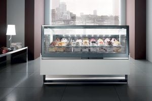 A modern gelato display case filled with various colorful gelato flavors sits in a sleek, contemporary room with large windows and cityscape views in the background.
