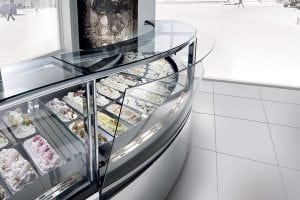 A modern gelato display case filled with various colorful gelato flavors, decorated with toppings, inside a bright, clean shop with large windows and tiled floors.