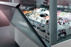 A display case filled with various colorful gelato and ice cream flavors, with the glass door open, showing different toppings and textures. The counter and surroundings are modern and clean.