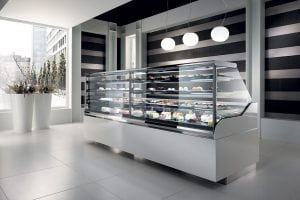 A modern bakery display case filled with assorted pastries and desserts stands in a sleek, contemporary shop with large windows, white floors, and three round hanging lights.