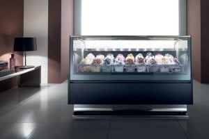 A modern ice cream display case with various colorful gelato flavors sits in a sleek, minimalistic room with tiled floors and a table holding a lamp and glassware.