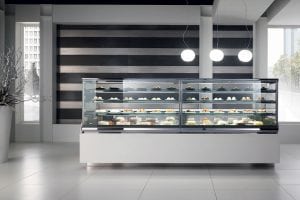 A modern bakery display case filled with assorted pastries and desserts stands in a sleek, minimalist shop with striped wall accents and two hanging round lights.