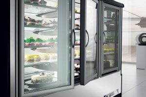 A display refrigerator with glass doors showing assorted cakes and pastries on multiple shelves in a modern, bright room with large windows.