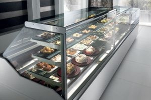 A glass display case in a modern bakery or café, filled with assorted pastries, cakes, and desserts arranged neatly on multiple shelves. The surroundings are bright and contemporary.