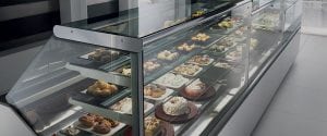 A modern glass display case filled with assorted pastries, cakes, and desserts on several shelves in a bright, clean bakery or café setting.