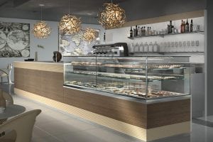 Modern café interior with a wood and glass display counter filled with pastries and desserts. Stylish pendant lights hang overhead, and shelves with bottles and glasses line the wall behind the counter.