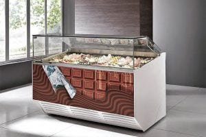 A modern ice cream display freezer with a glass top and a decorative front panel featuring a chocolate bar design, filled with various flavors of gelato in a bright, tiled room next to large windows.