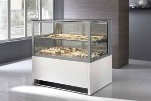 A modern glass display case filled with assorted pastries and baked goods stands in a clean, bright room with large windows and a gray accent wall.