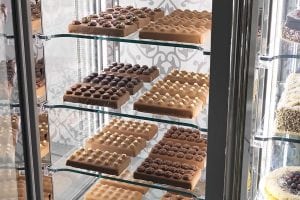 Rectangular trays of assorted chocolates and pralines are displayed on glass shelves inside a refrigerated display case, with decorative patterns visible on the back panel.