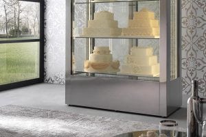A glass display case with three decorated white and cream wedding cakes stands in a modern room with patterned wallpaper, large window, and a light gray rug.