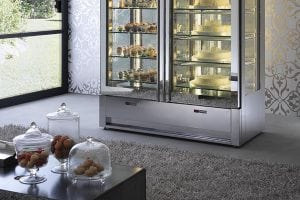 A modern room with a large glass display fridge filled with cakes and pastries. In the foreground, a dark table holds glass jars with assorted sweets. The room has patterned wallpaper and a large window.