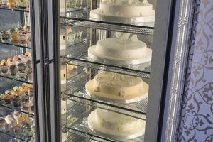A refrigerated display case with several white frosted cakes on glass shelves. The cakes have simple, elegant decorations. On the left, another case holds various cupcakes and desserts in glass cups.