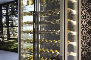 A modern glass wine fridge filled with bottles of wine, each sealed with a yellow cap, stands against a patterned wall. Large windows beside it reveal a green outdoor landscape with trees.