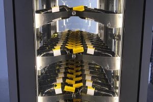Close-up view of a modern wine storage rack holding multiple rows of wine bottles placed horizontally, each with a yellow label, displayed behind glass and lit by vertical LED lights.