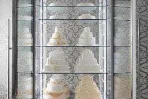 A display case with glass shelves shows several white tiered wedding cakes decorated with yellow and white floral designs, set against a patterned gray and white background.