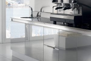 A modern kitchen with glossy, reflective cabinets, a sleek metal countertop, a sink with a faucet, and an espresso machine. Large windows reveal a view of water and boats outside.