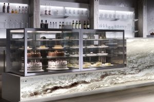 A modern bakery display counter with glass cases showcasing various cakes and pastries. Shelves behind hold bottles and glassware, and the counter front features a decorative marble-like design.