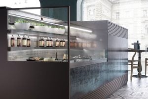 A modern self-service food counter with glass display, shelves of potted herbs, jars of condiments, and plates of food, set in a stylish café with large windows and city views.