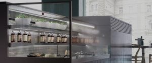 A modern glass display case in a cafe, showcasing bottles and potted herbs on shelves, with a cityscape visible through large windows in the background.