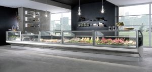 A modern deli or grocery store with large glass display cases filled with various meats, cheeses, and produce; contemporary decor, hanging lights, and large windows showing a cityscape outside.